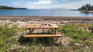 Une table de pique-nique en bois, ornée de quelques rochers, est installée sur une plage de sable, d’où l’on peut admirer l’océan et, au loin, une île couverte de forêts.