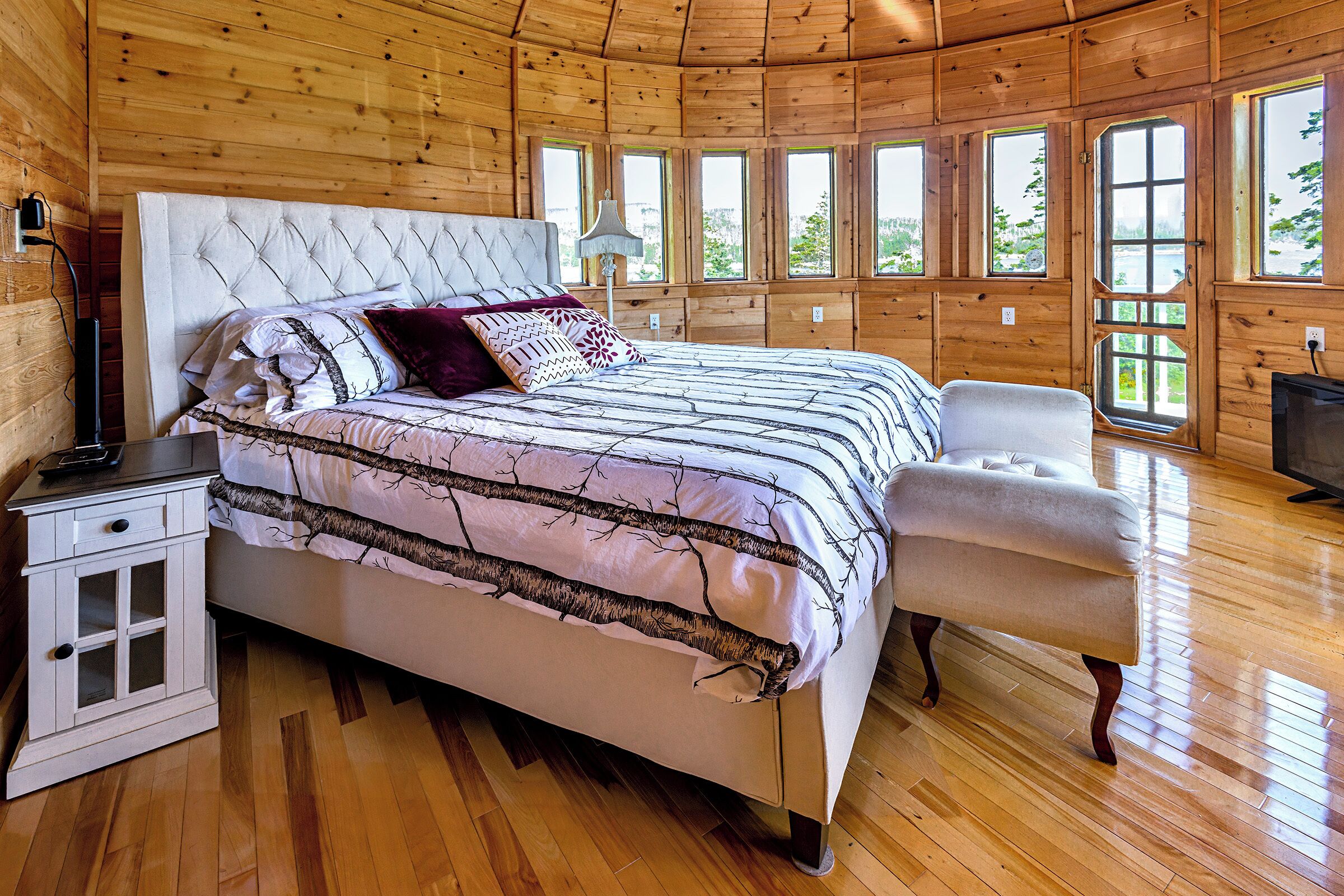 Un dormitorio de cabaña de madera con una cama grande, una mesita de noche, un banco y varias ventanas.