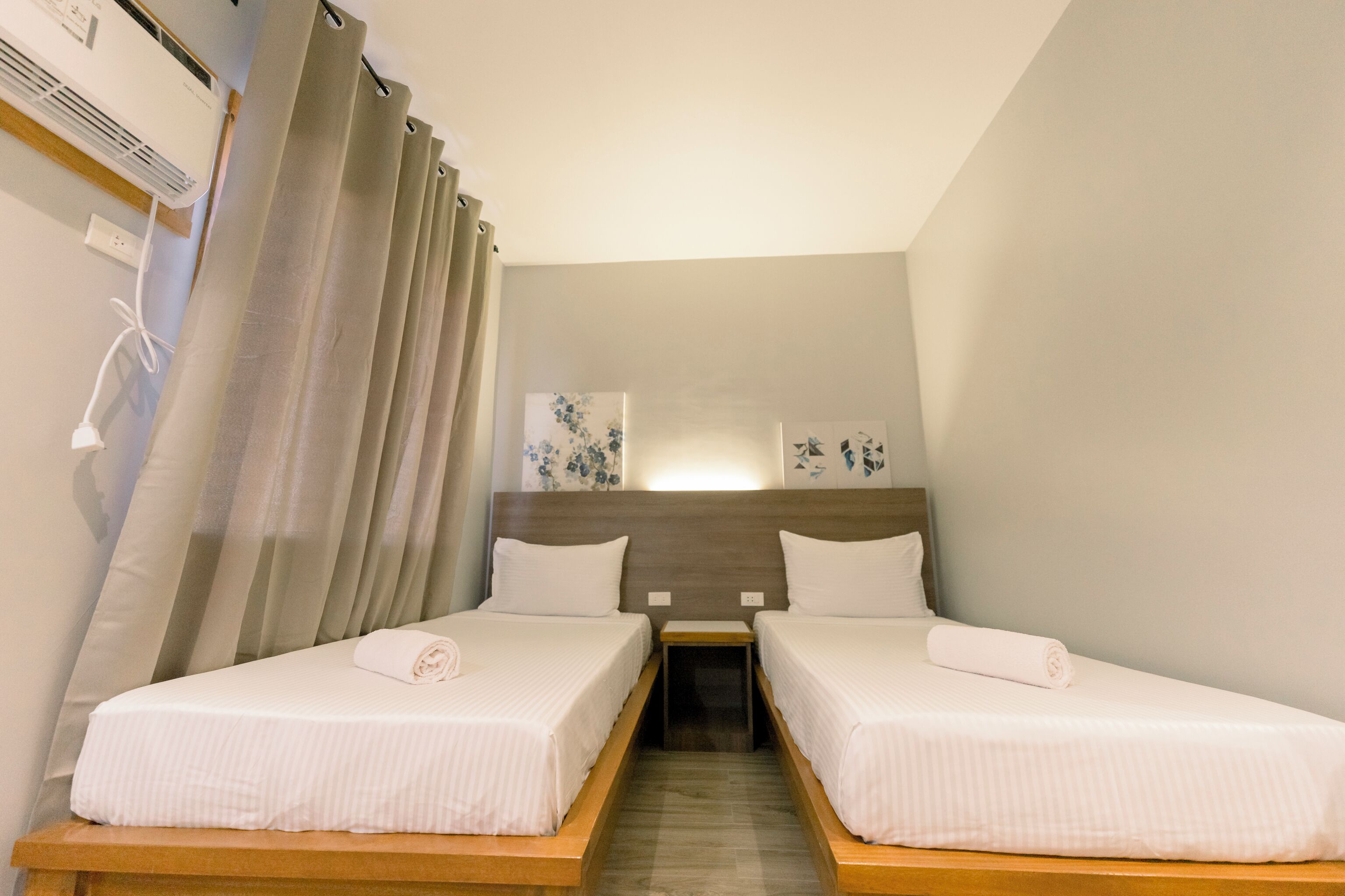 A hotel room with two single beds, a wooden nightstand, a wall-mounted air conditioner, and a curtain.