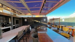 A modern outdoor dining area with a long wooden table and wicker chairs, a bar area with stools, and a large TV screen. - Urbani Smart Residence (João Pessoa)