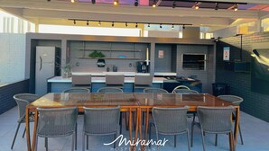 An outdoor kitchen area with a glass-top table, wicker chairs, and a built-in grill. - Urbani Smart Residence (João Pessoa)
