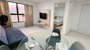 A modern hotel room with a sofa, a glass dining table with plates, a flat-screen TV mounted on the wall, and a bed with a white headboard. - Urbani Smart Residence (João Pessoa)