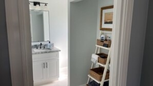 Un couloir mène à une salle de bain dotée d’un meuble-lavabo blanc et d’un miroir. On y trouve une étagère blanche avec des paniers et un tableau encadré accroché au mur.