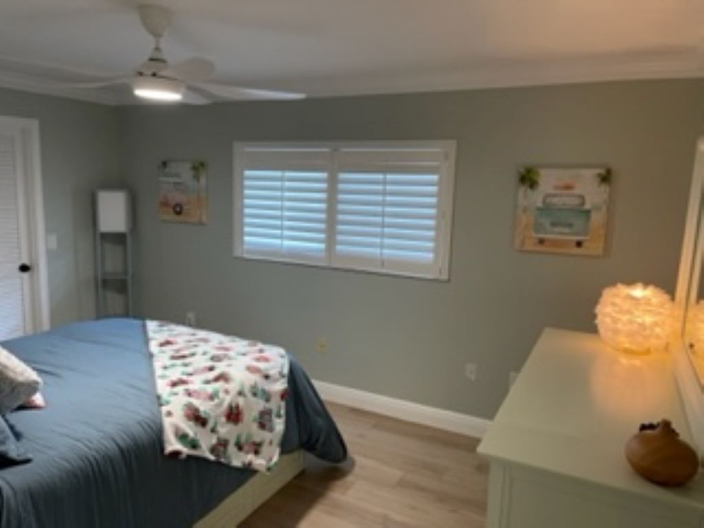 Quarto com cama, mesa de cabeceira, ventilador de teto, janela com persianas e quadros nas paredes.