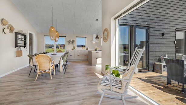 Un comedor amplio con una mesa larga y sillas, una cocina al fondo, y una terraza con una mecedora.
