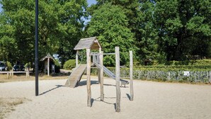 Un parque infantil con un tobogán de madera y una estructura para escalar, rodeado de arena y árboles.
