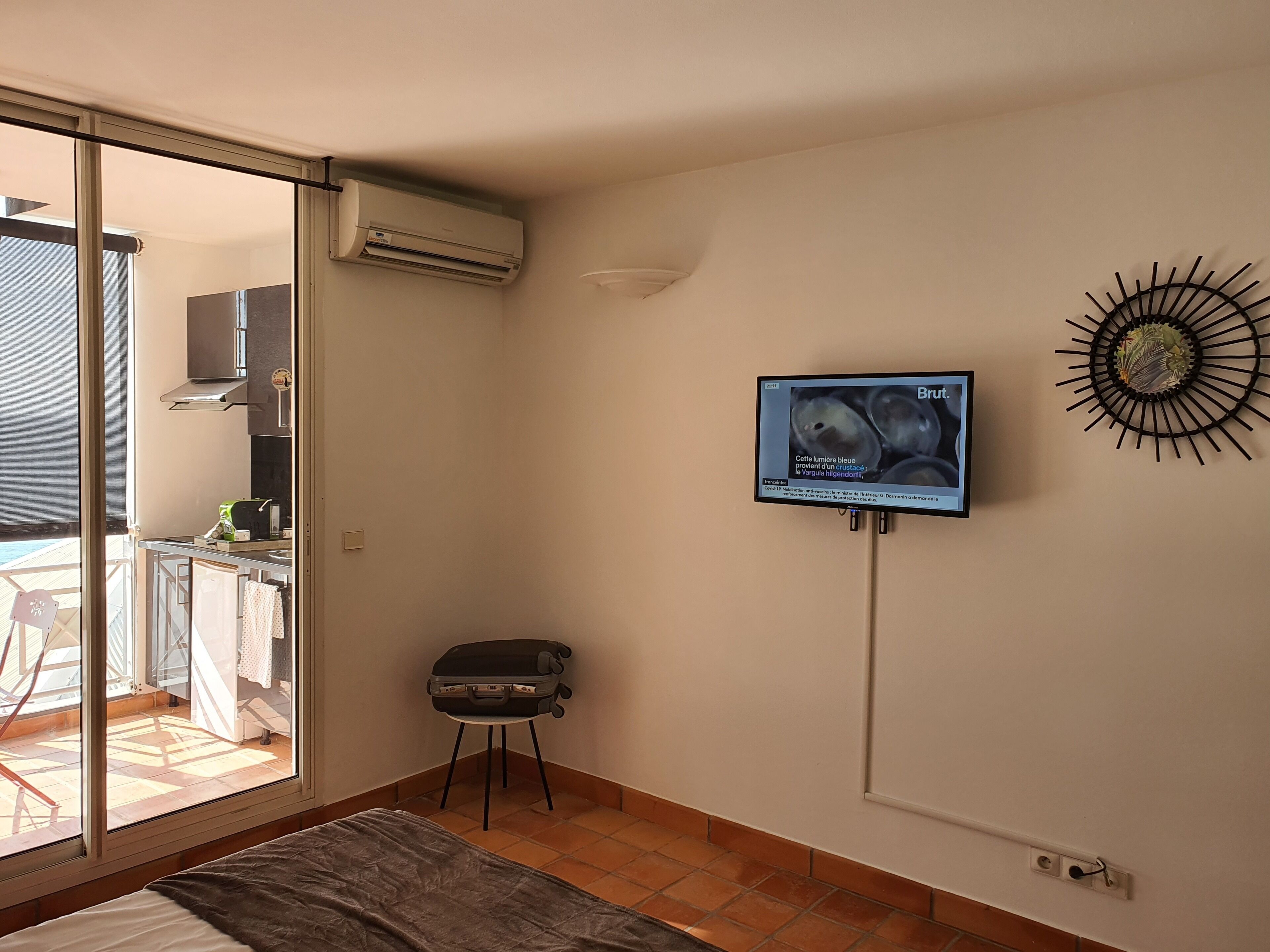 A bedroom with a bed, a suitcase, a television mounted on the wall, and a view of the balcony through a sliding door.