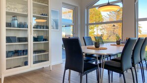 Un coin repas avec un buffet blanc où sont exposés des verres et des assiettes bleus, une table ronde avec des chaises, et de grandes fenêtres offrant une vue sur le feuillage automnal.