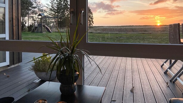 Une terrasse en bois avec une table et un pot de fleurs, donnant sur un champ verdoyant et un coucher de soleil.
