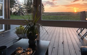 Eine Holzdielen-Terrasse mit Tisch und einer Topfpflanze, mit Blick auf ein Wiesenfeld und einen Sonnenuntergang.