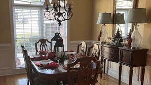 Une salle à manger avec une table en bois, des chaises et un lustre. On y trouve une fenêtre avec des stores, un miroir et un meuble avec des lampes.