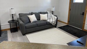 Una sala de estar con un sofá gris, dos almohadas blancas, una manta gris, dos mesitas de noche negras con lámparas, un tapete blanco y un control remoto negro sobre una mesa negra.