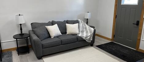 A living room with a grey sofa, two white pillows, a grey blanket, two black side tables with lamps, a white rug, and a black remote on a black table.