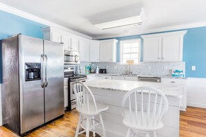 A modern kitchen with white cabinetry, a stainless steel refrigerator, a built-in oven, and a large island with two white chairs.