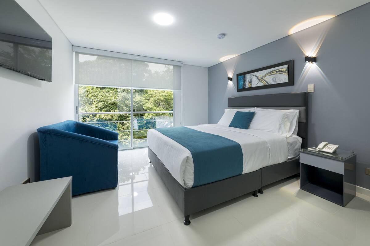 Un dormitorio moderno con una cama, una silla azul, una mesita de noche con un teléfono y un ventanal que ofrece vistas al verdor.