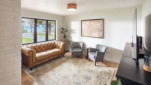 Ein Wohnzimmer mit einer Ledersofa, zwei Sesseln, einem Couchtisch und einer TV-Konsole. Ein großes Fenster bietet Blick auf eine Auffahrt und Bäume.