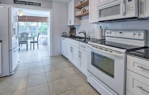 Una cocina moderna con armarios blancos, microondas integrado y estufa. Se ve un comedor con mesa y sillas a través de una puerta abierta.