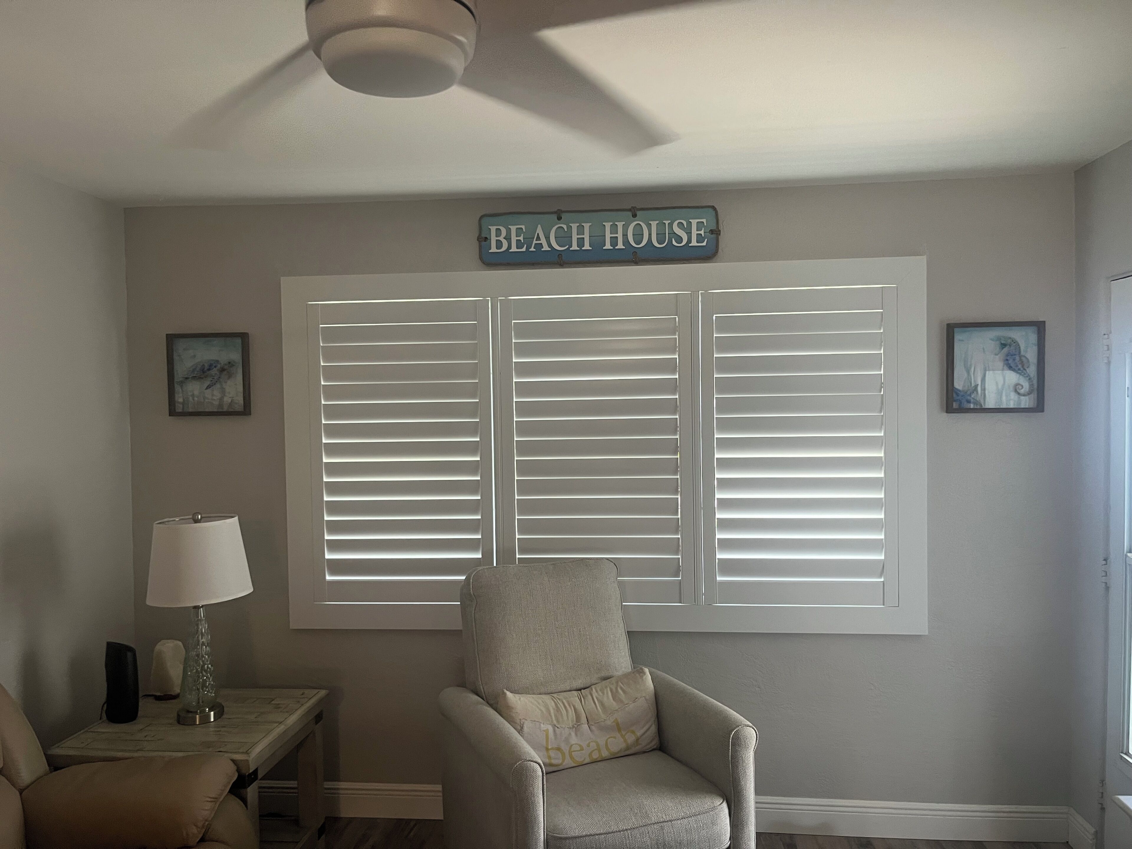 Ein Zimmer mit großem Fenster, weißen Fensterläden, einem Sessel, einer Lampe und einem Schild mit der Aufschrift 'BEACH HOUSE'.