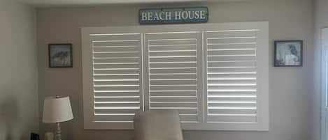 A room with a large window with white shutters, a chair, a lamp, and a sign that reads 'BEACH HOUSE'.