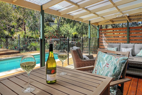 Uma área de estar coberta ao ar livre com piscina, uma garrafa de vinho e copos sobre uma mesa.