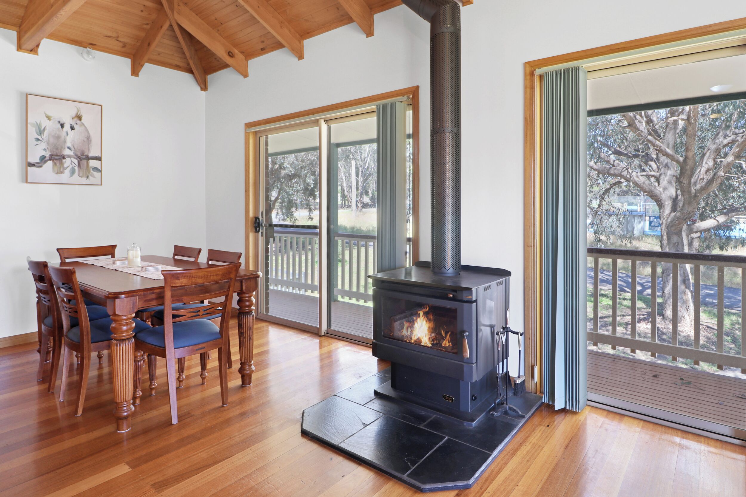 A room with a wooden dining table, chairs, a black wood-burning stove, and large windows with a view of trees.