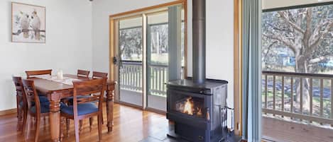 Una habitación con una mesa de comedor de madera, sillas, un estufa de leña negra y ventanas grandes con vistas a árboles.