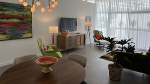 Ein modernes Wohnzimmer mit Essbereich, ausgestattet mit einem Kronleuchter, einem farbenfrohen abstrakten Gemälde, einem Flachbildfernseher und großen Fenstern mit transparenten Vorhängen.