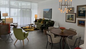 Ein modernes Wohnzimmer mit Essbereich, ausgestattet mit einem großen Fenster mit leichtem Vorhang, einer Kristallleuchtre und mehreren gerahmten Kunstwerken an den Wänden.