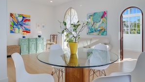 A modern dining area with a glass table, white chairs, a yellow vase with flowers, and abstract paintings on the walls. - Casa Estufa 25 (Funchal)