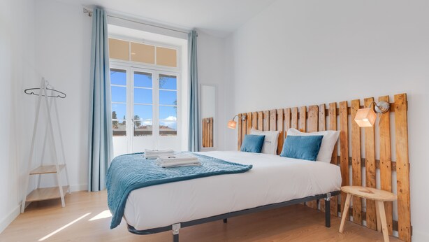 A bedroom with a wooden headboard, a bed with white bedding, a blue throw blanket, and a small wooden stool. - Casa Estufa 25 (Funchal)