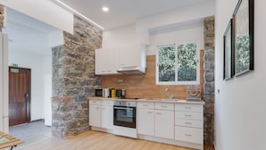 A modern kitchen with white cabinets, a stone accent wall, a built-in oven, and a window with a view of greenery. - Casa Estufa 25 (Funchal)
