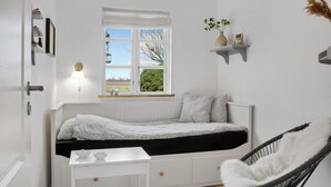 Un dormitorio minimalista con una cama blanca, una mesita de noche pequeña, una silla y una ventana con vistas a árboles.