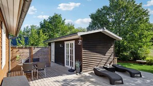 Una terraza de madera con mobiliario exterior, un pequeño edificio de madera y un área cercada con vegetación.