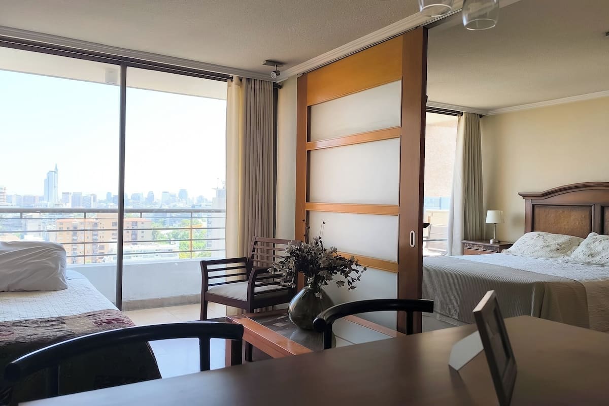 Quarto de hotel com cama, escrivaninha e cadeira. Vê-se uma vista da cidade através de grandes janelas.