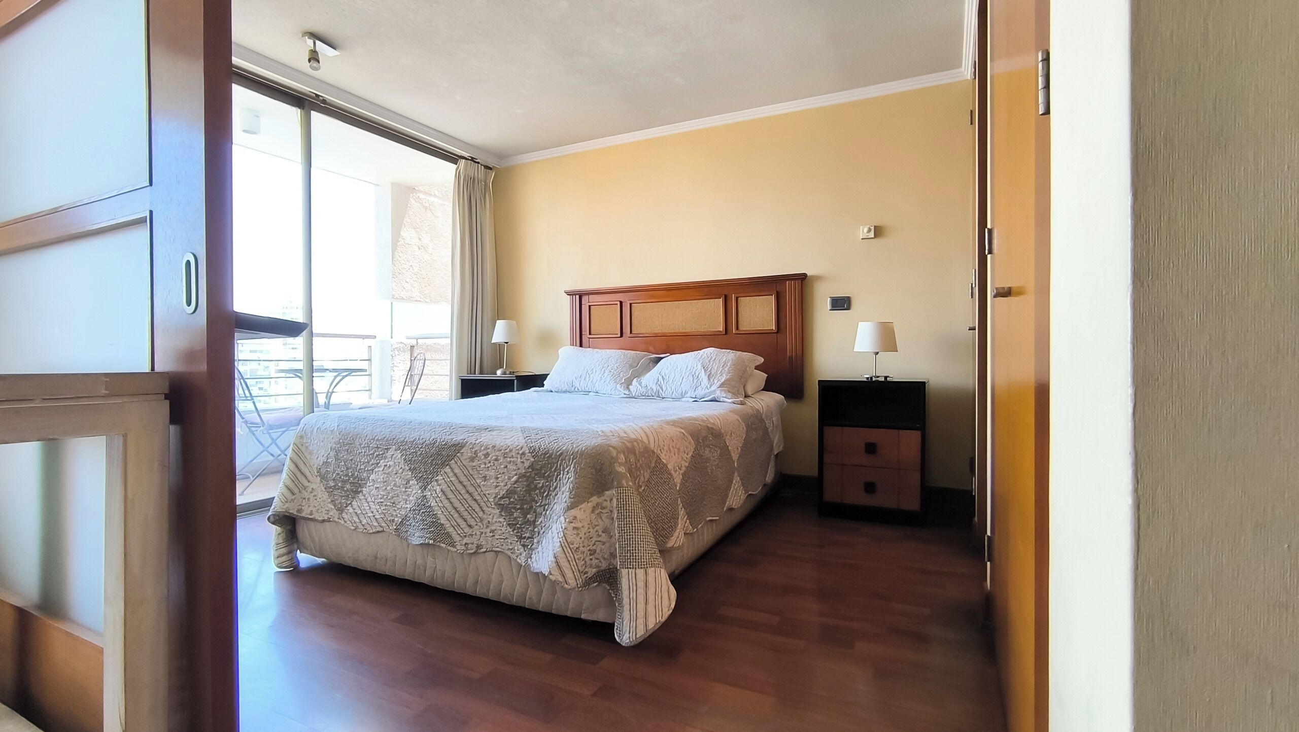 Une chambre à coucher avec un lit, une tête de lit en bois, deux tables de chevet avec des lampes, une porte-fenêtre coulissante donnant sur un balcon et un plancher en bois.