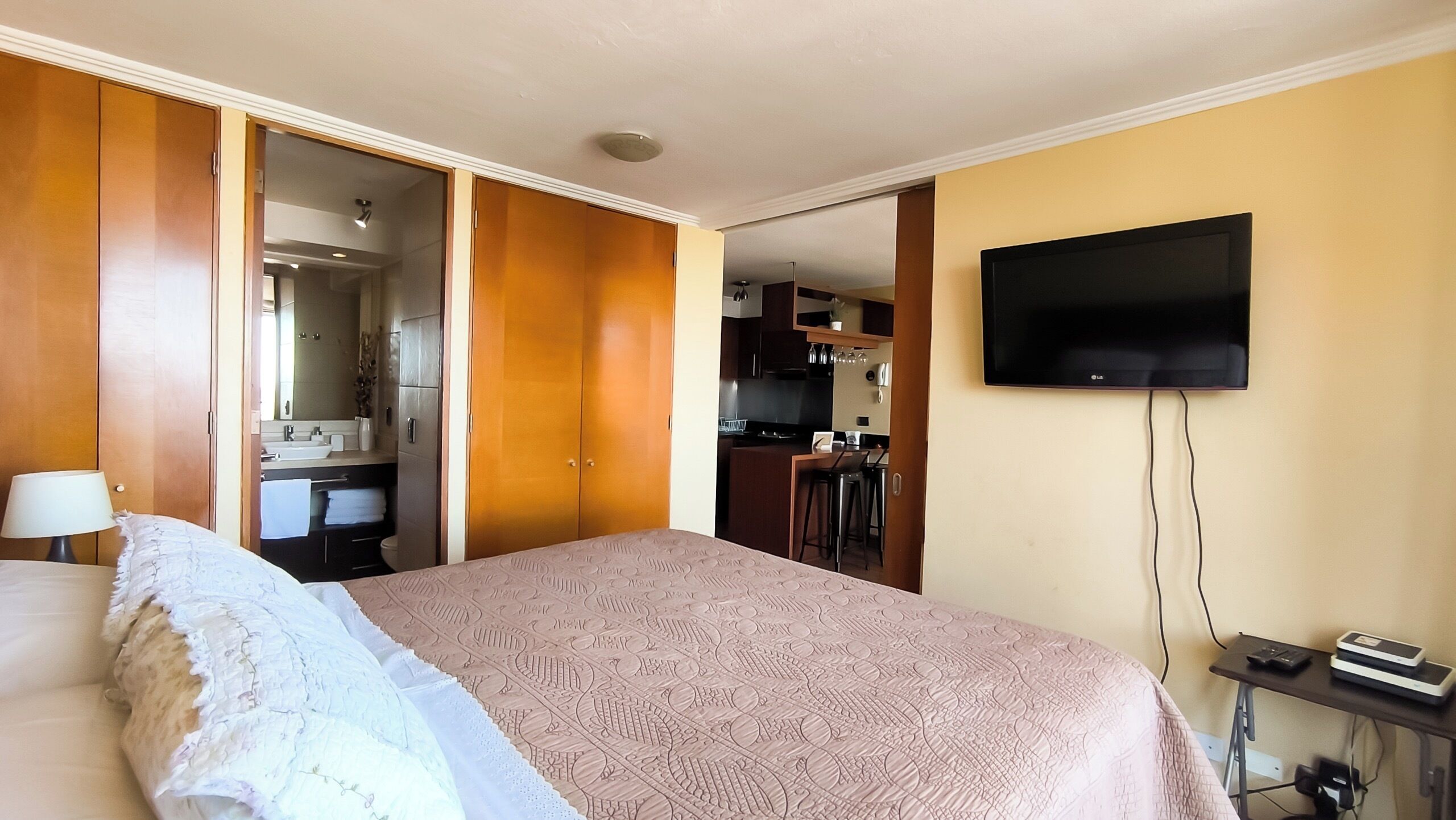 A hotel room with a bed, a TV, a desk with a computer, and a kitchenette in the background.