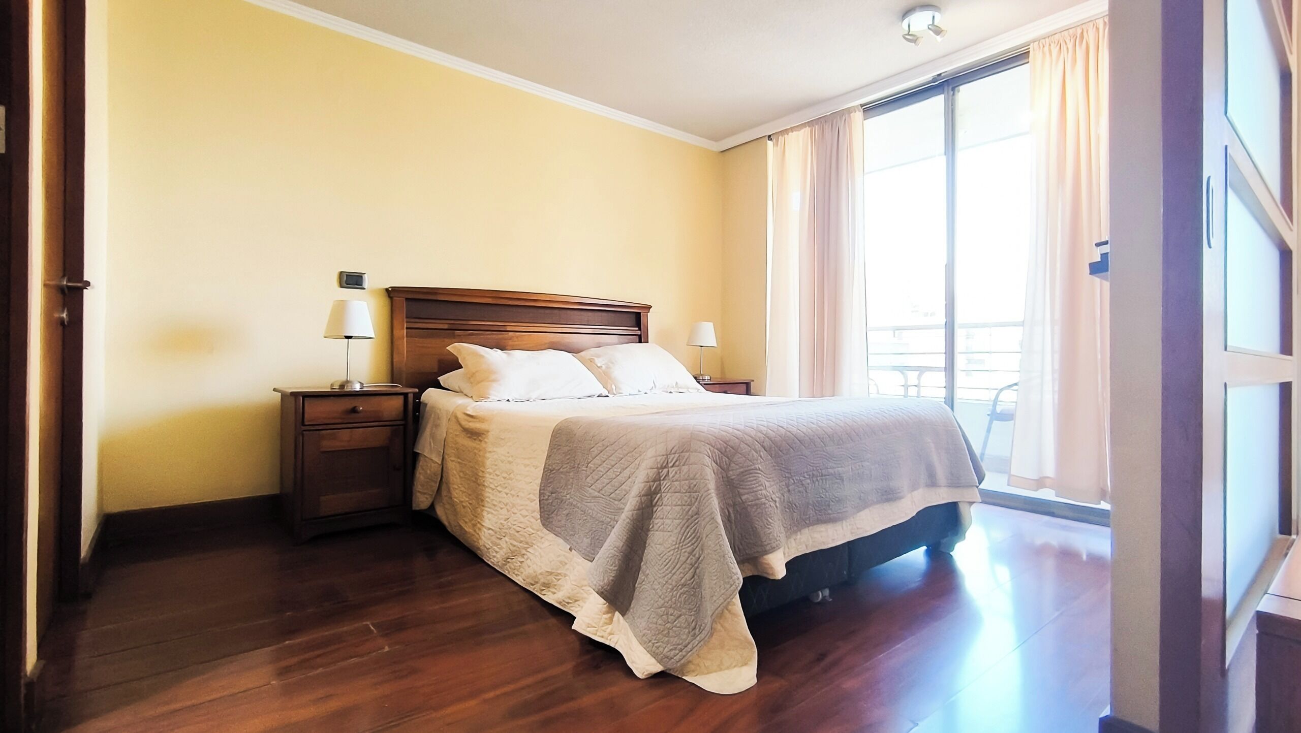Une chambre d’hôtel avec un lit en bois, deux tables de chevet avec des lampes, une grande fenêtre avec des rideaux et une vue sur l’extérieur.