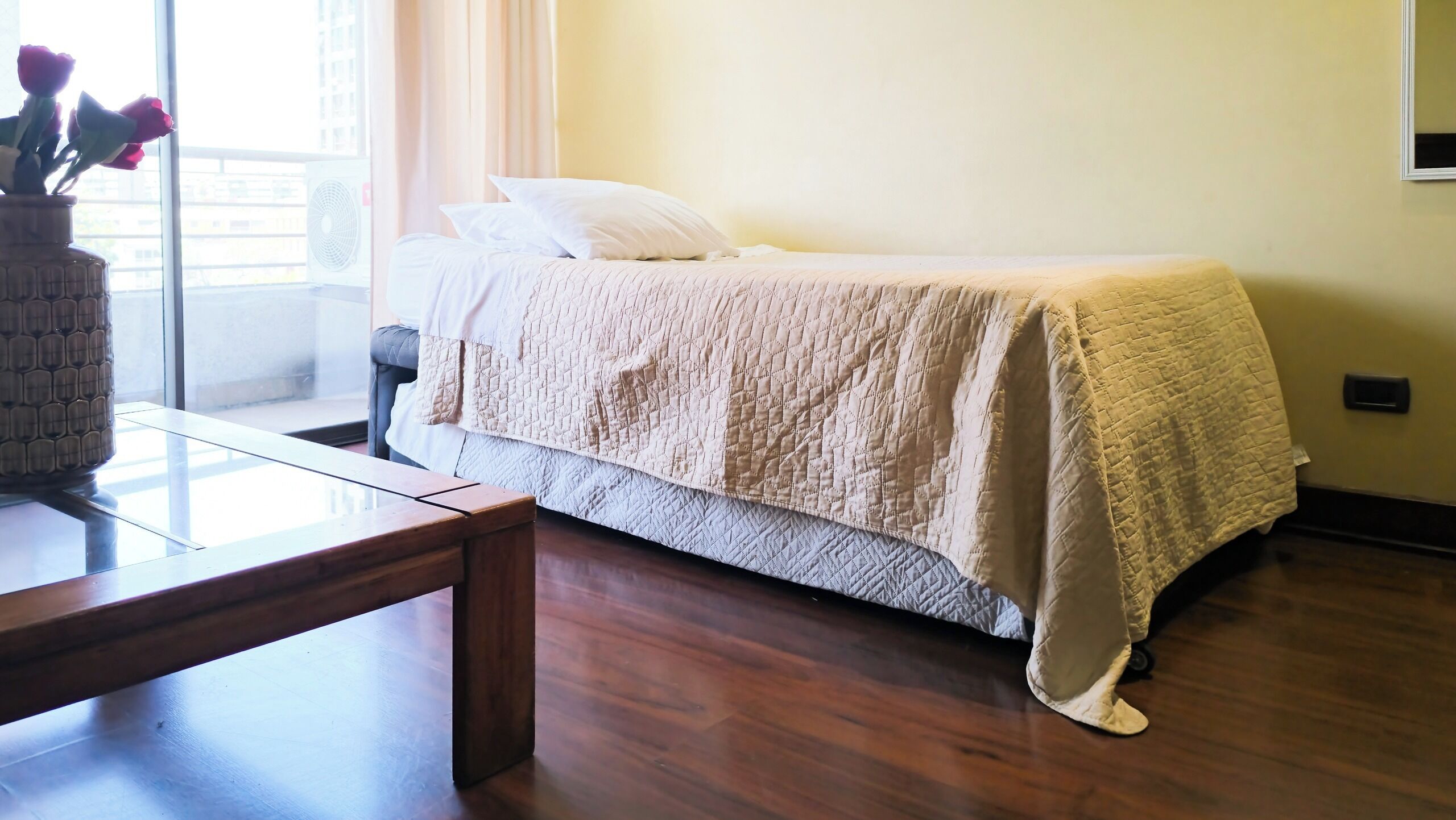 A neatly made bed with a quilted cover, a bedside table, and a vase with flowers.