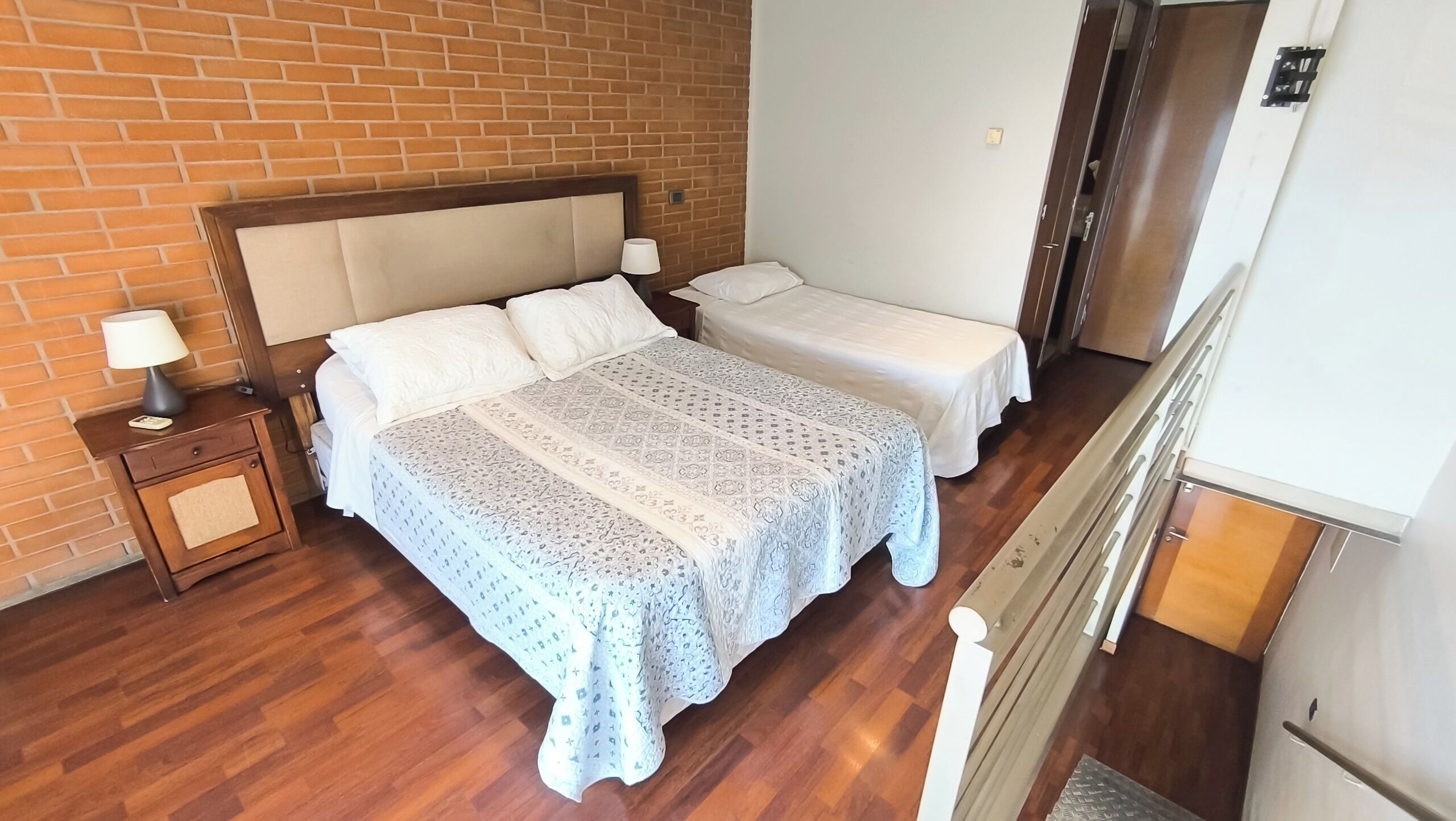 A hotel room with a brick wall, a large bed with a patterned bedspread, two bedside tables with lamps, and a wooden floor.