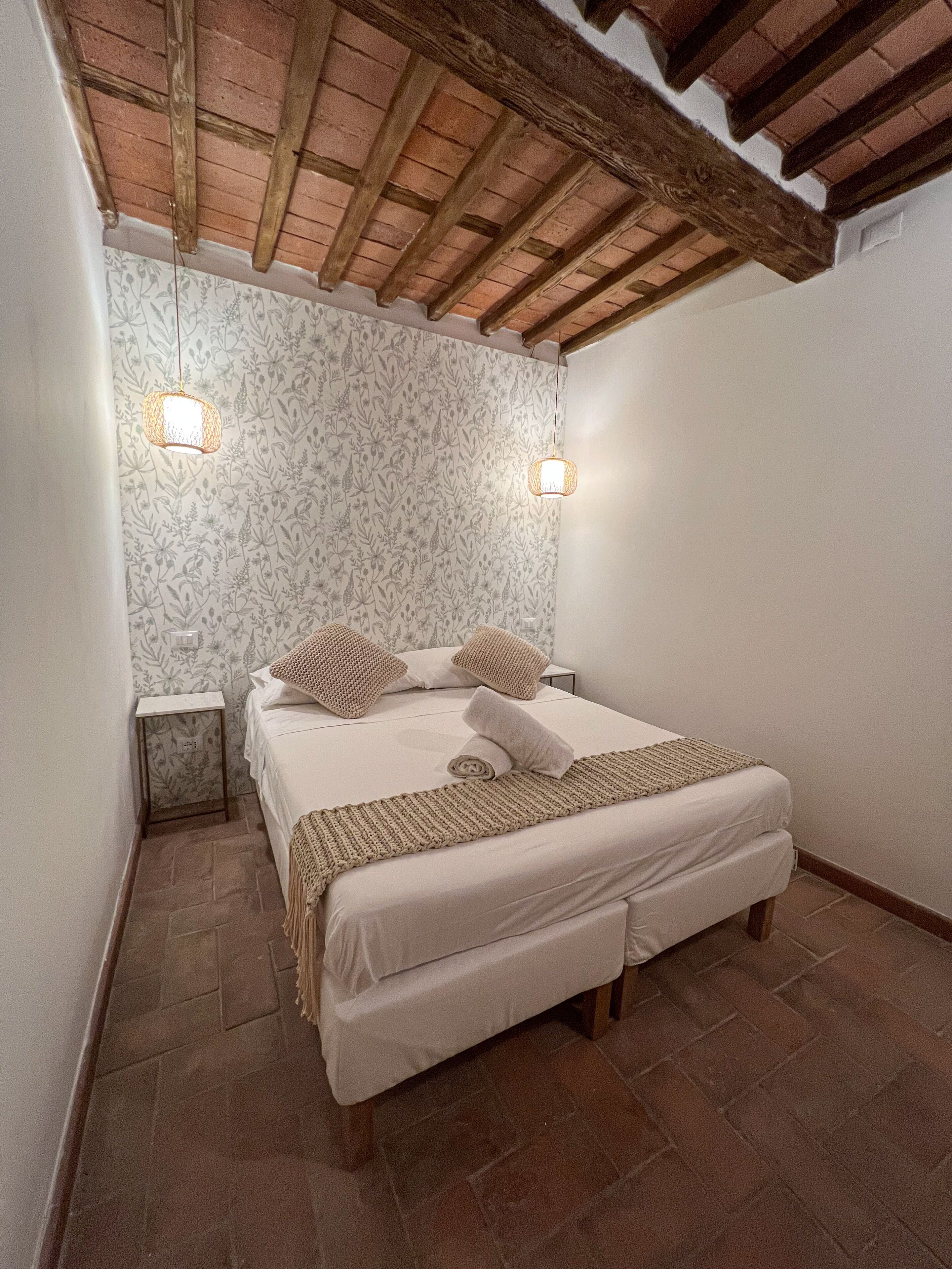 Une chambre avec un plafond en bois, un lit recouvert de draps blancs, deux lampes de chevet et un papier peint à motifs.