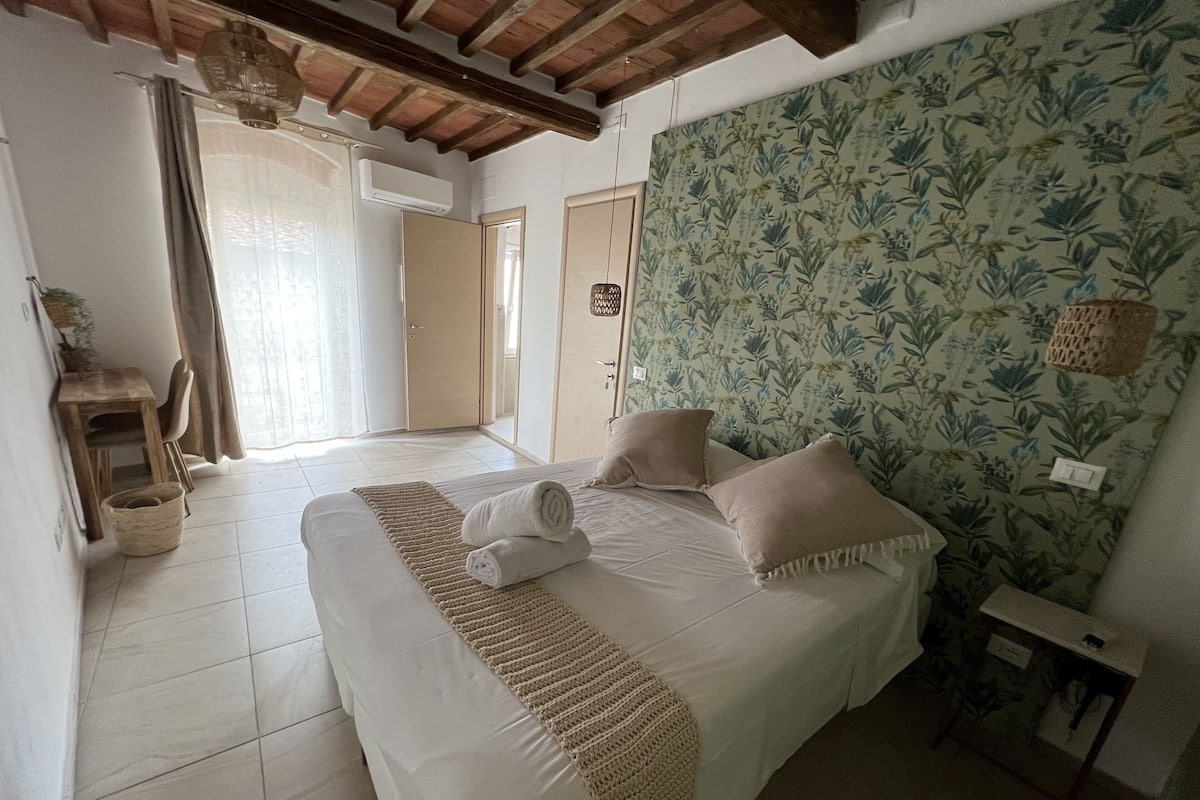A bedroom with a bed, a bedside table, a wall with floral wallpaper, a door, and a window with curtains.