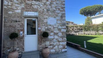 A stone building with a white door, two potted plants, and a sign that reads "INGRESSO - ENTRANCE".