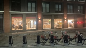 Une rangée de vélos est stationnée devant un immeuble qui abrite plusieurs commerces, dont l’un est reconnaissable à son enseigne lumineuse au néon.