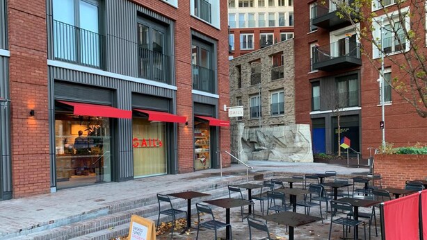 Un espace extérieur aménagé avec des tables et des chaises, un trottoir et un bâtiment doté d’une marquise rouge et d’une enseigne sur laquelle on peut lire « GAIL’S ».