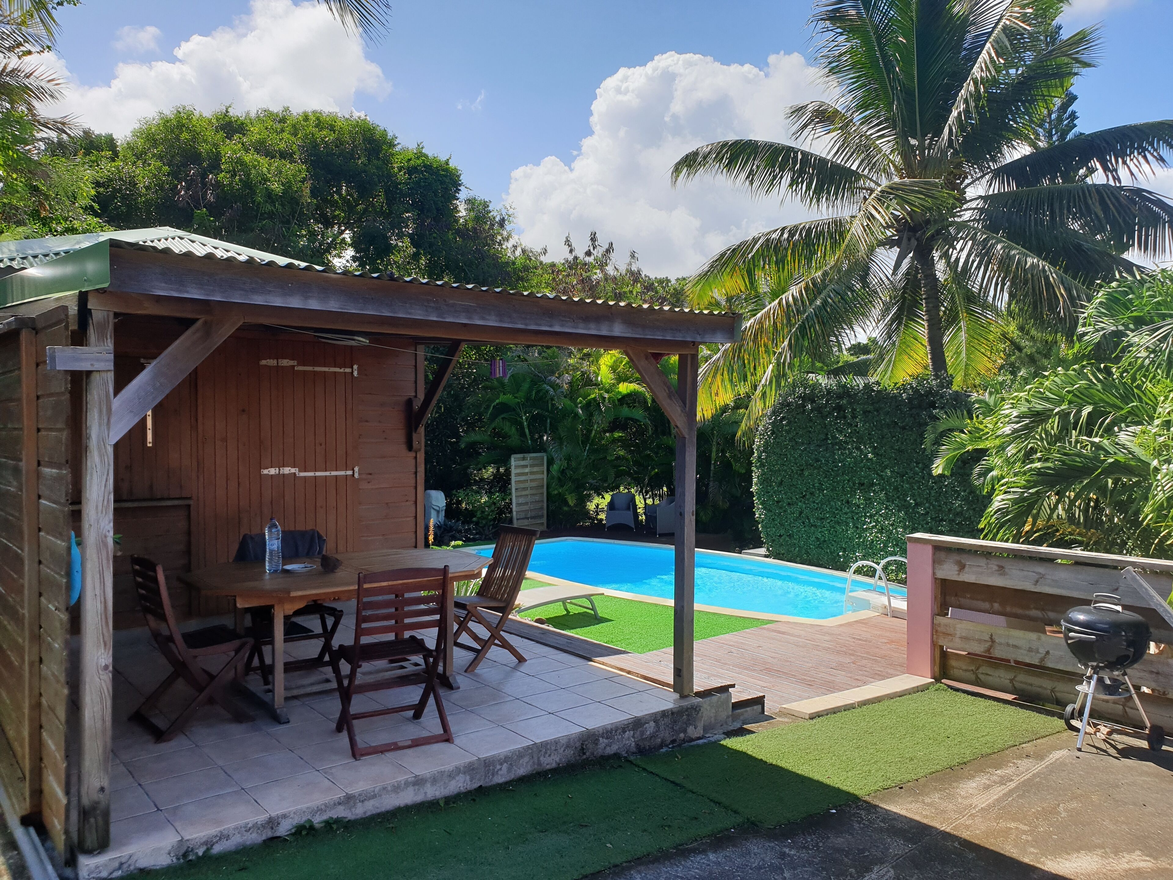 Un espacio de estar exterior con madera, que incluye una mesa, sillas, una parrilla y una piscina al fondo.