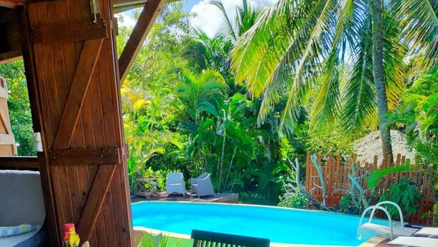 Una mesa de madera con un ramo de flores y una botella, frente a un fondo de piscina y exuberante vegetación.