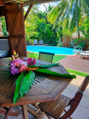 A wooden table with a bouquet of flowers and a bottle, set against a backdrop of a swimming pool and lush greenery. - COUP DE COEUR: charming wooden bungalow with private pool (Le Moule)