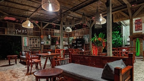 A rustic wooden interior with multiple seating areas, including benches and tables, under a thatched roof with hanging light fixtures. - Bali Jungle Camping (Pupuan)