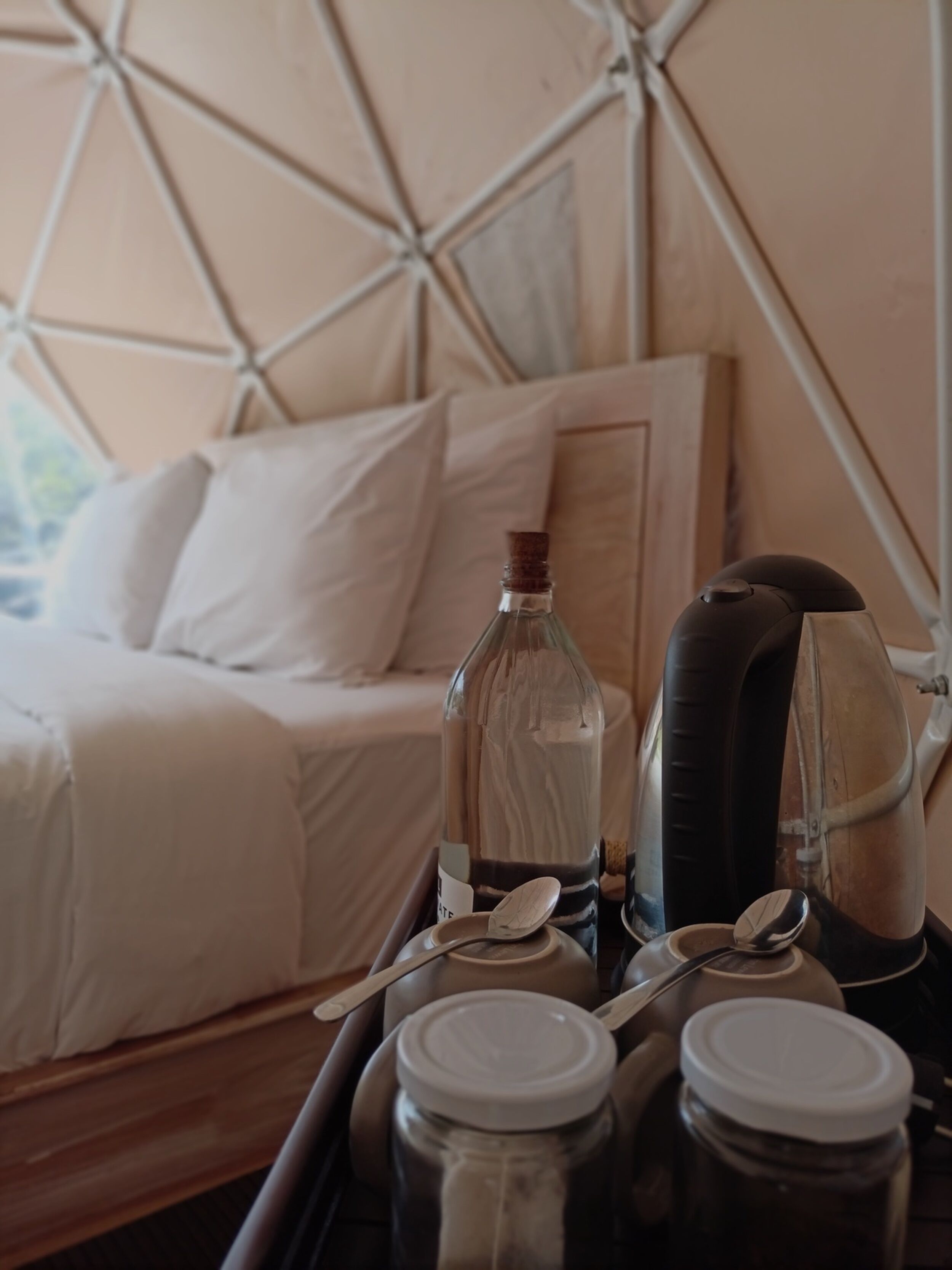 A bed with white bedding, a glass bottle, a kettle, and two jars on a tray.