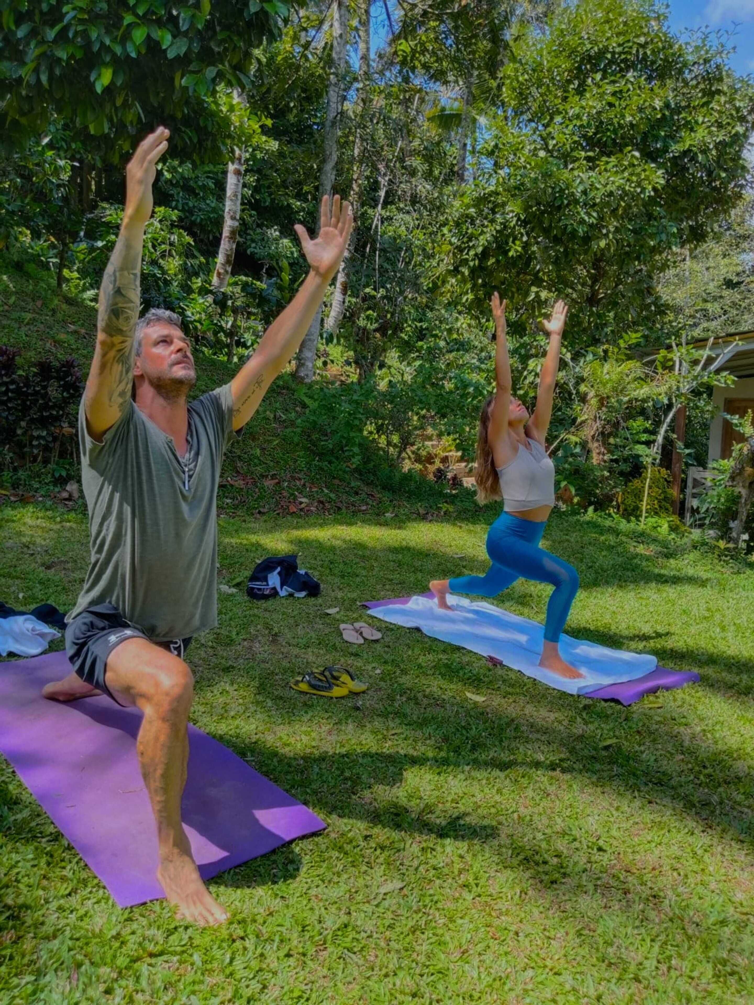 Two people practicing yoga on mats outdoors, surrounded by greenery.